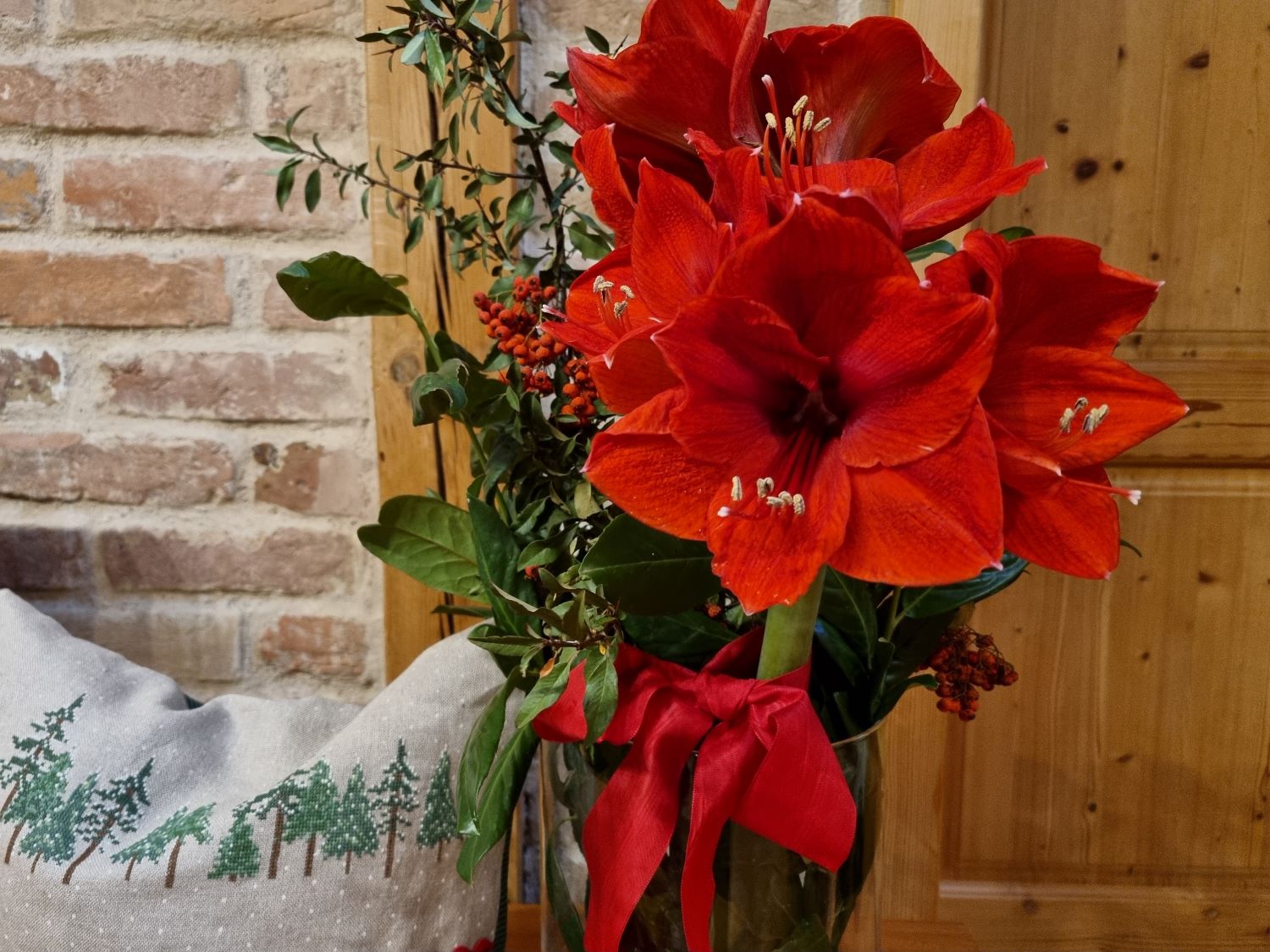 Eine Vase mit zwei vollaufgebluehten roten Amarilys, vor einer Ziegelmauer. ein kleines Kissen mit bestickten Baeumen.